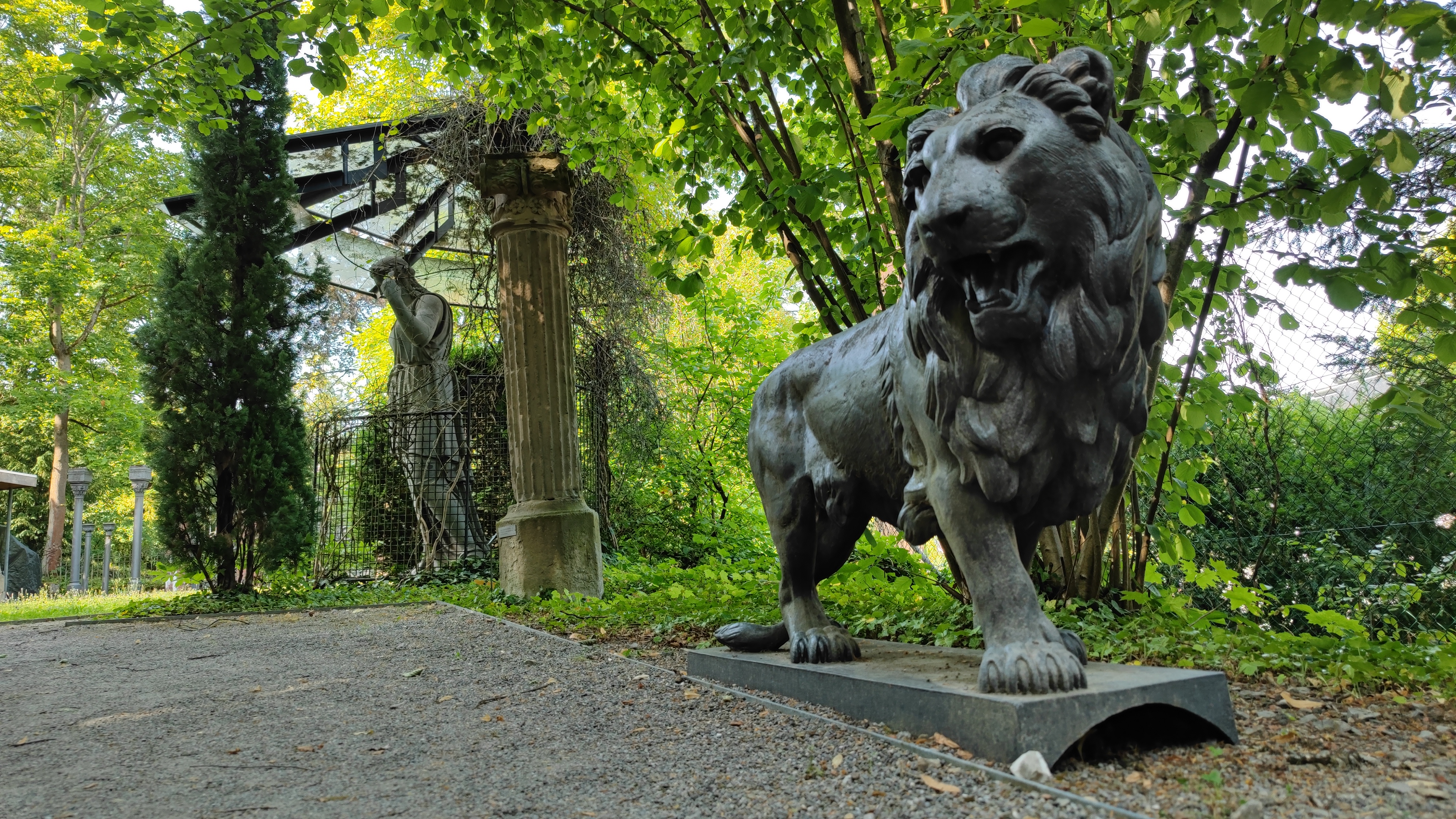 A statue of a lion up close, and another statue in the background, both along the gravel pathway.
